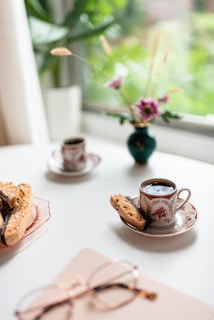 Elegant tea cups filled with dark coffee are placed on saucers on a white surface. A piece of biscotti rests beside one of the cups. In the background, a small green vase holds purple flowers, with greenery visible through a nearby window. Nearby, a pair of eyeglasses rests on a book or notebook.