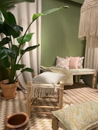 The image features a cozy interior setting with a predominantly green theme. A large potted plant with broad leaves adds a natural element. The seating area includes a wooden stool with a fringed cushion and a wooden bench adorned with floral and patterned cushions. A woven rug covers the floor, complementing the earthy tones of the decor. A decorative hanging piece with tassels adds texture near the ceiling.