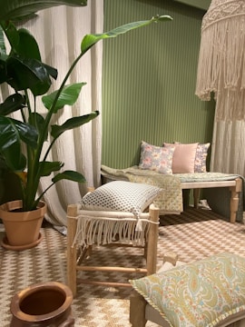 The image features a cozy interior setting with a predominantly green theme. A large potted plant with broad leaves adds a natural element. The seating area includes a wooden stool with a fringed cushion and a wooden bench adorned with floral and patterned cushions. A woven rug covers the floor, complementing the earthy tones of the decor. A decorative hanging piece with tassels adds texture near the ceiling.