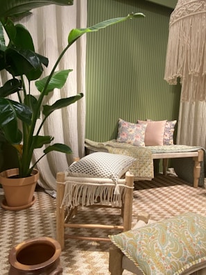 The image features a cozy interior setting with a predominantly green theme. A large potted plant with broad leaves adds a natural element. The seating area includes a wooden stool with a fringed cushion and a wooden bench adorned with floral and patterned cushions. A woven rug covers the floor, complementing the earthy tones of the decor. A decorative hanging piece with tassels adds texture near the ceiling.