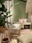 The image features a cozy interior setting with a predominantly green theme. A large potted plant with broad leaves adds a natural element. The seating area includes a wooden stool with a fringed cushion and a wooden bench adorned with floral and patterned cushions. A woven rug covers the floor, complementing the earthy tones of the decor. A decorative hanging piece with tassels adds texture near the ceiling.