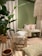 The image features a cozy interior setting with a predominantly green theme. A large potted plant with broad leaves adds a natural element. The seating area includes a wooden stool with a fringed cushion and a wooden bench adorned with floral and patterned cushions. A woven rug covers the floor, complementing the earthy tones of the decor. A decorative hanging piece with tassels adds texture near the ceiling.