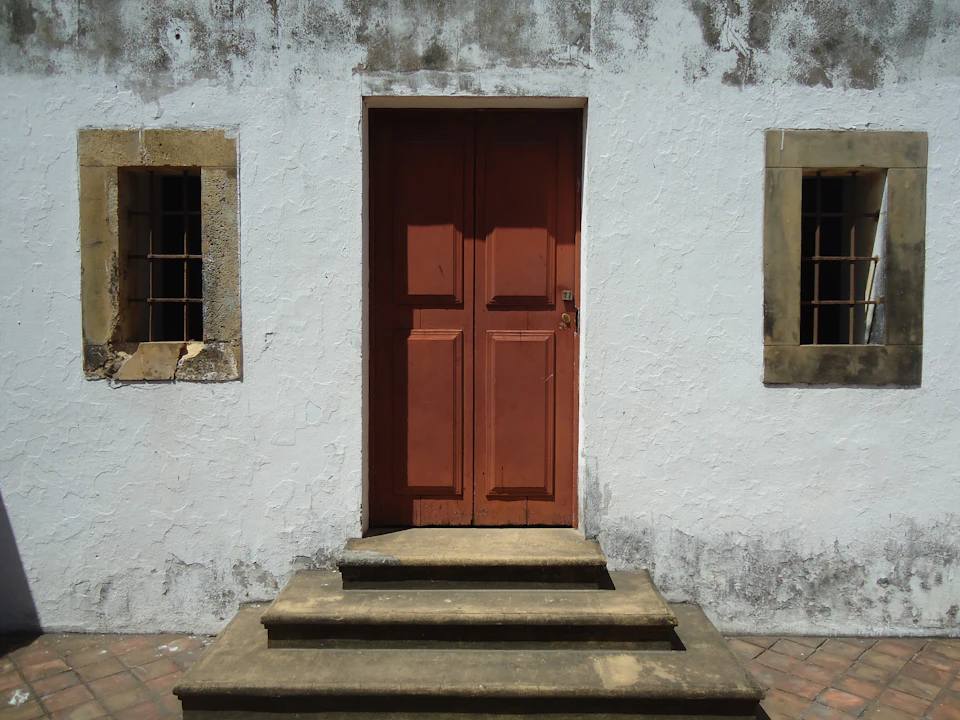 The image shows a white stone building with two windows, one on each side of the door. The door is painted red and features a wooden handle. A set of stairs leads up to the door, which is located at the bottom right corner of the frame. The building appears to be in good condition, with no visible signs of damage or wear.