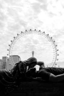 A black and white photograph featuring a couple in an intimate pose with a large Ferris wheel in the background. One person is laying down, while the other leans over, their faces close together. The cityscape and architectural elements provide a modern setting, with the wheel towering in the distance.