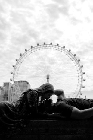 A black and white photograph featuring a couple in an intimate pose with a large Ferris wheel in the background. One person is laying down, while the other leans over, their faces close together. The cityscape and architectural elements provide a modern setting, with the wheel towering in the distance.