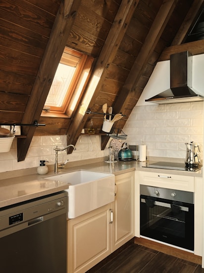 A cozy kitchen with wooden slanted ceilings, featuring a farmhouse sink, modern stainless steel appliances, and a white tile backsplash. Sunlight streams through a skylight, illuminating the space. Kitchen utensils and a kettle are neatly placed on the countertop, adding to the warm, inviting atmosphere.