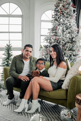 A family of three sitting on a green couch in a festive setting with a Christmas tree in the background. The man and woman are seated next to a young boy holding a teddy bear. The room has large arched windows and is decorated with holiday ornaments.