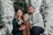 A family of three, consisting of a man, woman, and a child, is posing together amidst festive, snowy Christmas trees. They are smiling and appear to be enjoying a holiday setting, with the child holding a brown teddy bear. The background includes white and green decorated trees, glowing lights, and a wooden ladder.