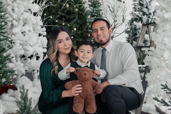A family of three, consisting of a man, woman, and a child, is posing together amidst festive, snowy Christmas trees. They are smiling and appear to be enjoying a holiday setting, with the child holding a brown teddy bear. The background includes white and green decorated trees, glowing lights, and a wooden ladder.