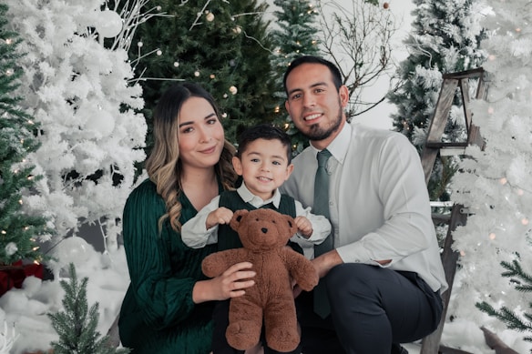 A family of three, consisting of a man, woman, and a child, is posing together amidst festive, snowy Christmas trees. They are smiling and appear to be enjoying a holiday setting, with the child holding a brown teddy bear. The background includes white and green decorated trees, glowing lights, and a wooden ladder.