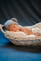 A sleeping baby wearing a knitted gray hat with bear ears, resting on a soft white blanket in a wicker basket. The background is a deep blue, creating a serene and peaceful atmosphere.