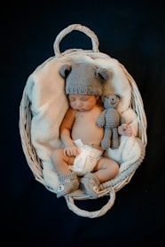 A baby is sleeping in a white wicker basket, wrapped in a soft blanket. The baby is wearing a knitted hat with bear ears, matching booties, and a diaper. There is also a small knitted bear beside the baby.