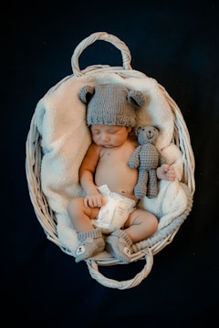A baby is sleeping in a white wicker basket, wrapped in a soft blanket. The baby is wearing a knitted hat with bear ears, matching booties, and a diaper. There is also a small knitted bear beside the baby.