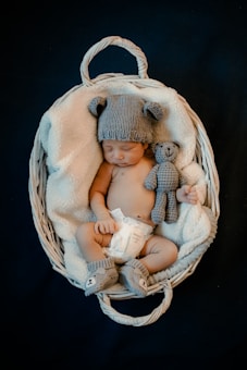 A baby is sleeping in a white wicker basket, wrapped in a soft blanket. The baby is wearing a knitted hat with bear ears, matching booties, and a diaper. There is also a small knitted bear beside the baby.