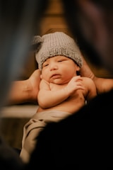 A newborn baby is carefully held, swaddled in a soft blanket. The baby wears a cozy knitted hat and has a serene, sleepy expression.