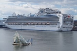 A large cruise ship docked at a port, displaying multiple decks and life rafts. The ship is white with blue accents and has the lettering 'Princess Cruises' on the side. In the foreground, an artistic glass structure floats on the water, resembling broken shards or ice.