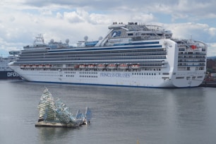 A large cruise ship docked at a port, displaying multiple decks and life rafts. The ship is white with blue accents and has the lettering 'Princess Cruises' on the side. In the foreground, an artistic glass structure floats on the water, resembling broken shards or ice.