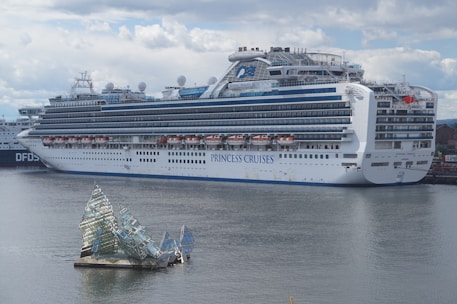 A large cruise ship docked at a port, displaying multiple decks and life rafts. The ship is white with blue accents and has the lettering 'Princess Cruises' on the side. In the foreground, an artistic glass structure floats on the water, resembling broken shards or ice.