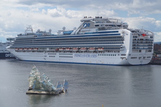 A large cruise ship docked at a port, displaying multiple decks and life rafts. The ship is white with blue accents and has the lettering 'Princess Cruises' on the side. In the foreground, an artistic glass structure floats on the water, resembling broken shards or ice.