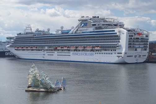 A large cruise ship docked at a port, displaying multiple decks and life rafts. The ship is white with blue accents and has the lettering 'Princess Cruises' on the side. In the foreground, an artistic glass structure floats on the water, resembling broken shards or ice.
