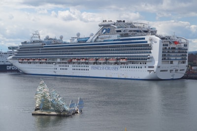 A large cruise ship docked at a port, displaying multiple decks and life rafts. The ship is white with blue accents and has the lettering 'Princess Cruises' on the side. In the foreground, an artistic glass structure floats on the water, resembling broken shards or ice.