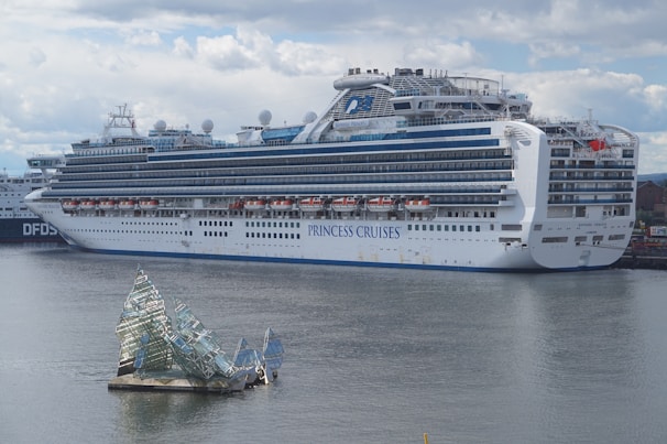 A large cruise ship docked at a port, displaying multiple decks and life rafts. The ship is white with blue accents and has the lettering 'Princess Cruises' on the side. In the foreground, an artistic glass structure floats on the water, resembling broken shards or ice.