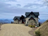 A picturesque house with stone and wood elements sits atop a hill, surrounded by a vast, natural landscape. The architecture is rustic and charming, featuring a gravel driveway leading up to the main entrance. In the background, a range of distant mountains is visible under a cloudy sky, enhancing the serene and remote setting.