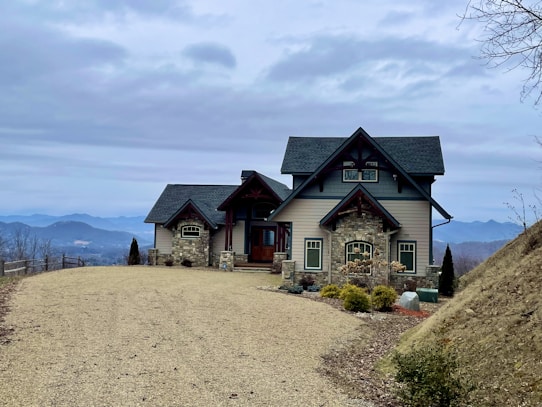 A picturesque house with stone and wood elements sits atop a hill, surrounded by a vast, natural landscape. The architecture is rustic and charming, featuring a gravel driveway leading up to the main entrance. In the background, a range of distant mountains is visible under a cloudy sky, enhancing the serene and remote setting.