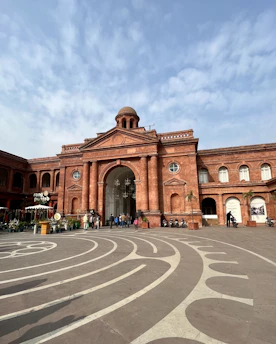 A grand red-brick building with a central dome and large arched entrance. The facade features classical architecture with columns and circular windows. The foreground includes a patterned stone plaza with curved designs. People are walking near the entrance, and there is a sign for a place named 'Rang Punjabi.' Several motorcycles are parked nearby.