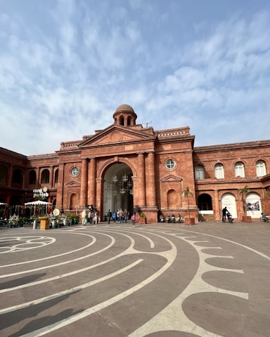 A grand red-brick building with a central dome and large arched entrance. The facade features classical architecture with columns and circular windows. The foreground includes a patterned stone plaza with curved designs. People are walking near the entrance, and there is a sign for a place named 'Rang Punjabi.' Several motorcycles are parked nearby.