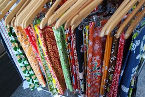 A collection of colorful patterned fabrics hanging on wooden hangers, featuring intricate designs with floral and geometric patterns in a variety of colors including orange, green, red, and blue.