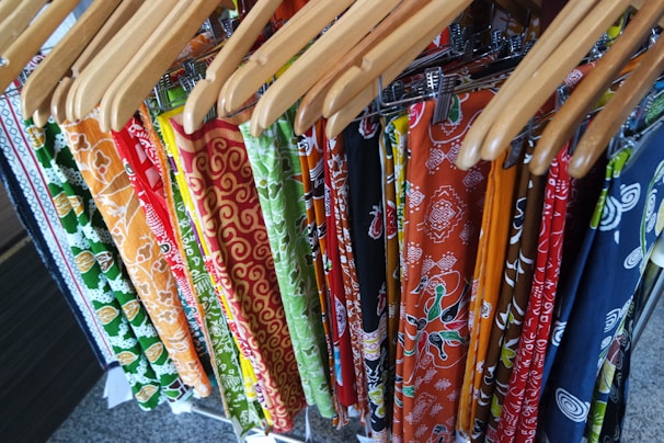 A collection of colorful patterned fabrics hanging on wooden hangers, featuring intricate designs with floral and geometric patterns in a variety of colors including orange, green, red, and blue.