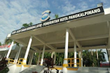 A pavilion with a large roof supported by white columns is prominently displayed. It has a sign on top that reads ‘Alun-Alun Taman Merdeka (ATM) Pemerintah Kota Pangkalpinang’. Surrounding the structure are palm trees and greenery, and the weather appears sunny with a partly cloudy sky.
