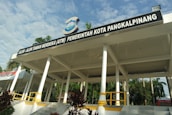 A pavilion with a large roof supported by white columns is prominently displayed. It has a sign on top that reads ‘Alun-Alun Taman Merdeka (ATM) Pemerintah Kota Pangkalpinang’. Surrounding the structure are palm trees and greenery, and the weather appears sunny with a partly cloudy sky.