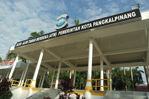 A pavilion with a large roof supported by white columns is prominently displayed. It has a sign on top that reads ‘Alun-Alun Taman Merdeka (ATM) Pemerintah Kota Pangkalpinang’. Surrounding the structure are palm trees and greenery, and the weather appears sunny with a partly cloudy sky.