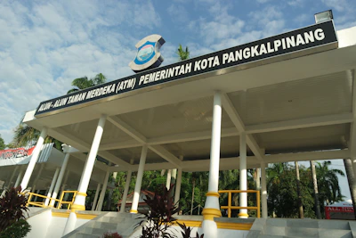 A pavilion with a large roof supported by white columns is prominently displayed. It has a sign on top that reads ‘Alun-Alun Taman Merdeka (ATM) Pemerintah Kota Pangkalpinang’. Surrounding the structure are palm trees and greenery, and the weather appears sunny with a partly cloudy sky.