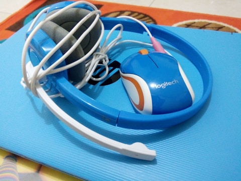 A blue Logitech computer mouse is positioned on a soft blue surface. Beside it is a pair of tangled wired headphones, also blue, with a white and gray microphone attached. The headphones and mouse are placed on what appears to be a laptop or notebook cover.