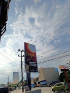 A large billboard stands prominently at a street intersection, displaying the word 'Surya' against a gradient background. Utility poles with numerous power lines intersect the view against a partly cloudy sky. Nearby, a few vehicles, including a green car, are visible along the road. Buildings with commercial signage line the street, contributing to an urban setting.