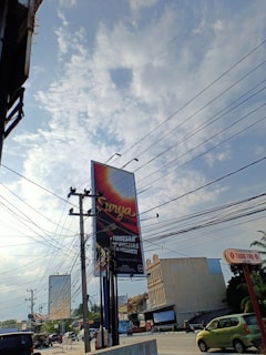 A large billboard stands prominently at a street intersection, displaying the word 'Surya' against a gradient background. Utility poles with numerous power lines intersect the view against a partly cloudy sky. Nearby, a few vehicles, including a green car, are visible along the road. Buildings with commercial signage line the street, contributing to an urban setting.