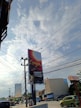 A large billboard stands prominently at a street intersection, displaying the word 'Surya' against a gradient background. Utility poles with numerous power lines intersect the view against a partly cloudy sky. Nearby, a few vehicles, including a green car, are visible along the road. Buildings with commercial signage line the street, contributing to an urban setting.