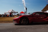A sleek, red sports car is prominently displayed in the foreground on a paved road. In the background, a group of people is gathered around a tent marked 'Top-End Motors,' with a second red canopy nearby. The setting seems to be an outdoor event with a clear blue sky and a grassy area.