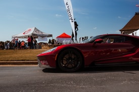 A sleek, red sports car is prominently displayed in the foreground on a paved road. In the background, a group of people is gathered around a tent marked 'Top-End Motors,' with a second red canopy nearby. The setting seems to be an outdoor event with a clear blue sky and a grassy area.