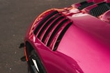 A close-up of a vibrant pink car detail, focusing on the sleek, aerodynamic vent on the car's body against a dark asphalt background. The curves and design suggest a modern, high-performance vehicle.