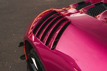 A close-up of a vibrant pink car detail, focusing on the sleek, aerodynamic vent on the car's body against a dark asphalt background. The curves and design suggest a modern, high-performance vehicle.