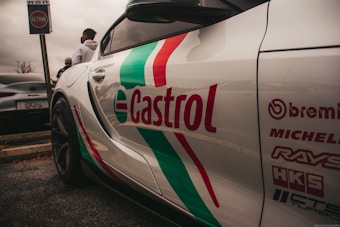 A sports car with prominent branding and logos on its side, including Castrol, Brembo, Michelin, Rays, HKS, and others, parked next to another car. The scene includes people standing nearby and a sign with 'Octane' in the background. The overall setting appears to be a parking lot on a cloudy day.