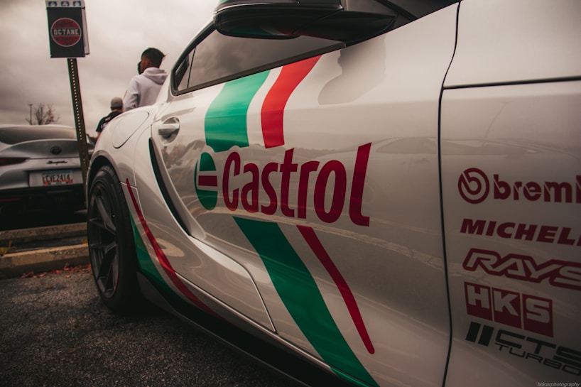 A sports car with prominent branding and logos on its side, including Castrol, Brembo, Michelin, Rays, HKS, and others, parked next to another car. The scene includes people standing nearby and a sign with 'Octane' in the background. The overall setting appears to be a parking lot on a cloudy day.