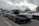 A sleek black sports car with a rooftop cargo box is parked in a lot filled with other vehicles. The car has tinted windows and a polished exterior, reflecting the overcast sky above. Other cars, including a silver sedan and a white vehicle, are visible nearby. People are gathered in the background, suggesting a car meet-up or show. Trees and poles line the parking area under cloudy skies.