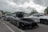 A sleek black sports car with a rooftop cargo box is parked in a lot filled with other vehicles. The car has tinted windows and a polished exterior, reflecting the overcast sky above. Other cars, including a silver sedan and a white vehicle, are visible nearby. People are gathered in the background, suggesting a car meet-up or show. Trees and poles line the parking area under cloudy skies.