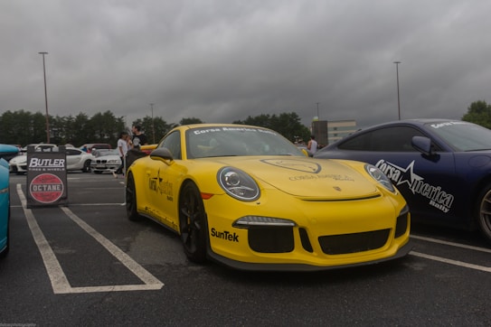 A yellow sports car with 'Corsa America Rally' branding is prominently displayed in a parking lot. Next to it is a blue car with similar branding. There are several people in the background, and a sign advertising tires and wheels is visible. The sky is overcast, adding a moody feel to the setting.