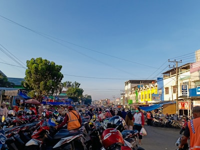 A bustling street scene featuring a large group of parked motorcycles, with people walking around and buildings lining the sides of the street. The sky is clear blue, and there are trees visible on the left side. Many people are wearing helmets and some are carrying goods, suggesting a market or busy area.